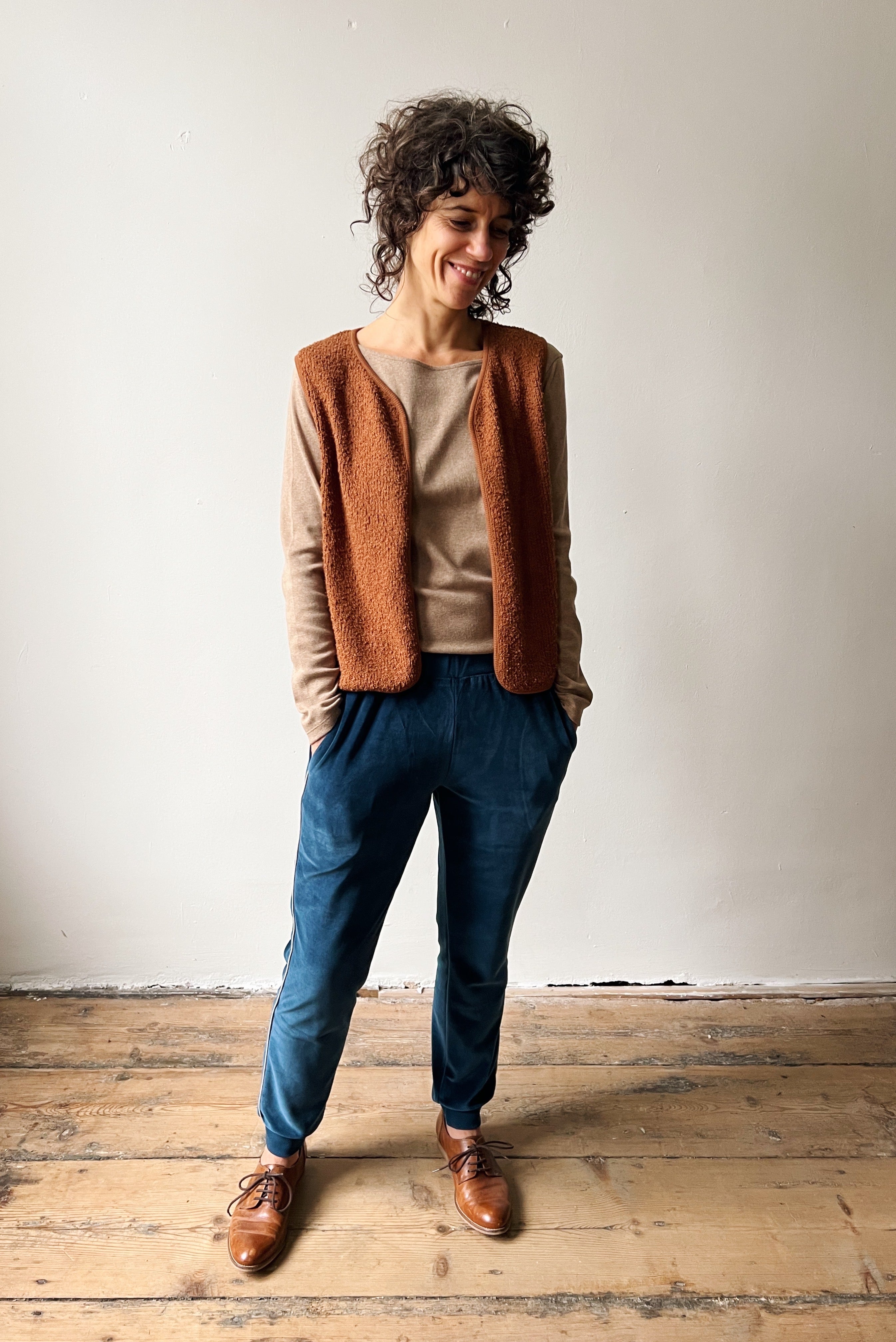 Woman wearing a brown vest, beige longsleeve, ocean blue sweatpants, and brown shoes standing on a wooden floor.