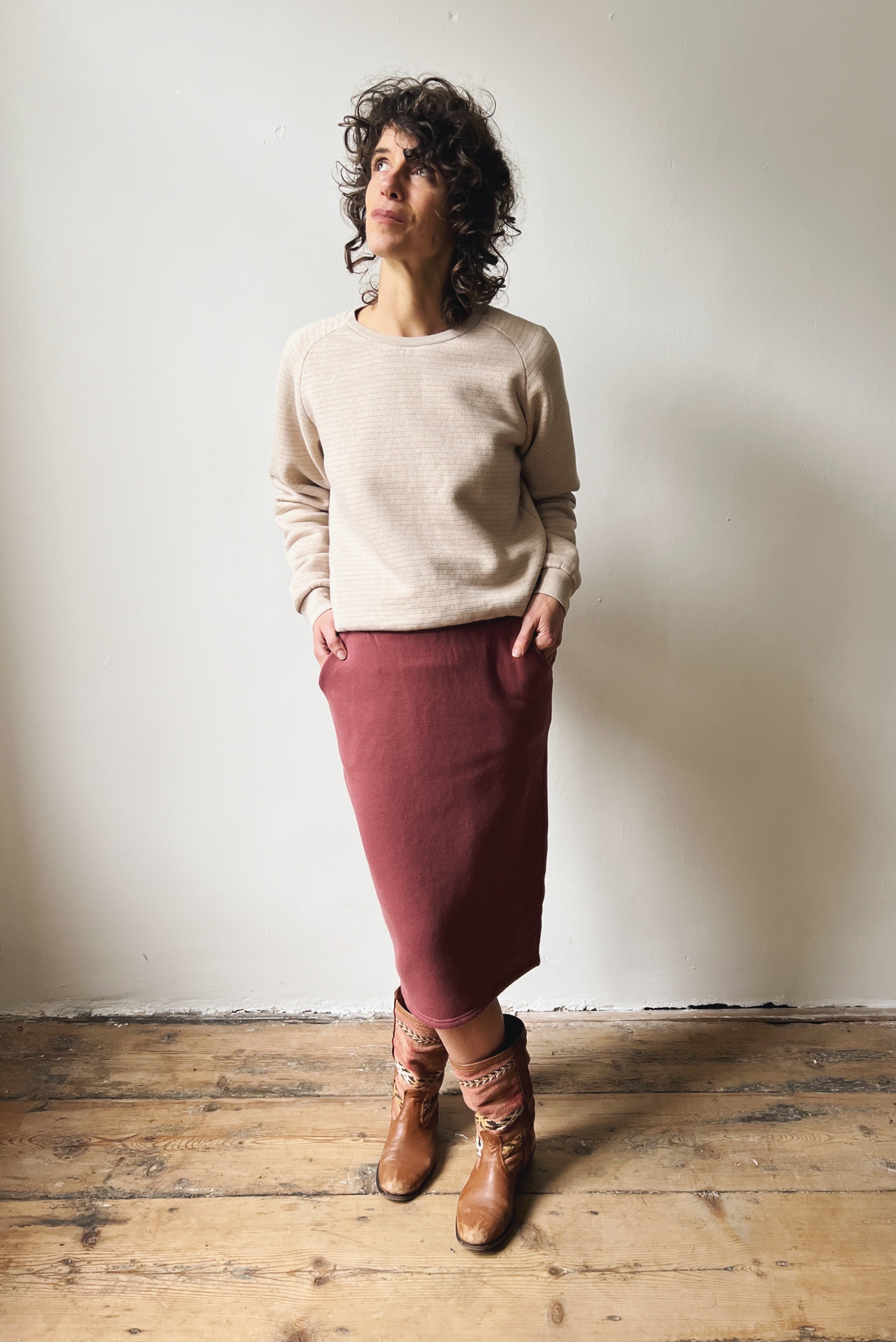 Woman wearing a beige sweater and figue skirt standing against a white wall.