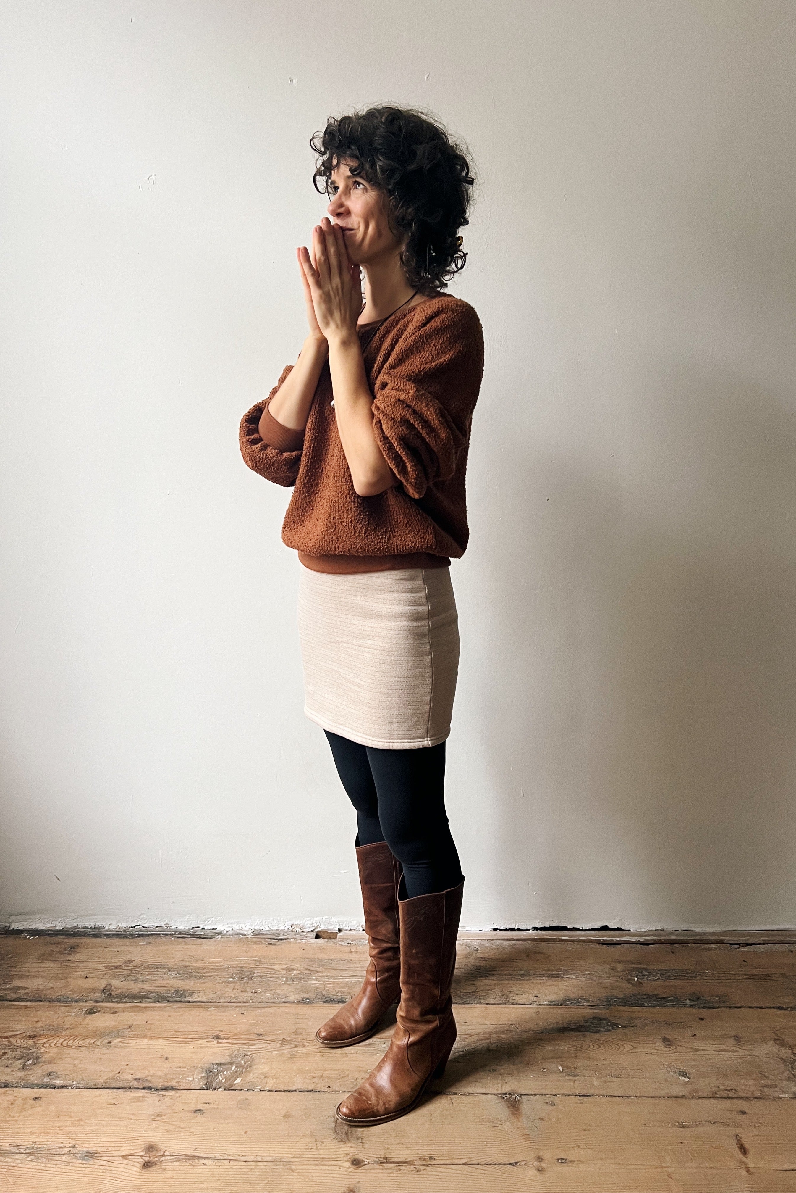 Woman wearing a brown sweater and beige skirt standing against a white wall.