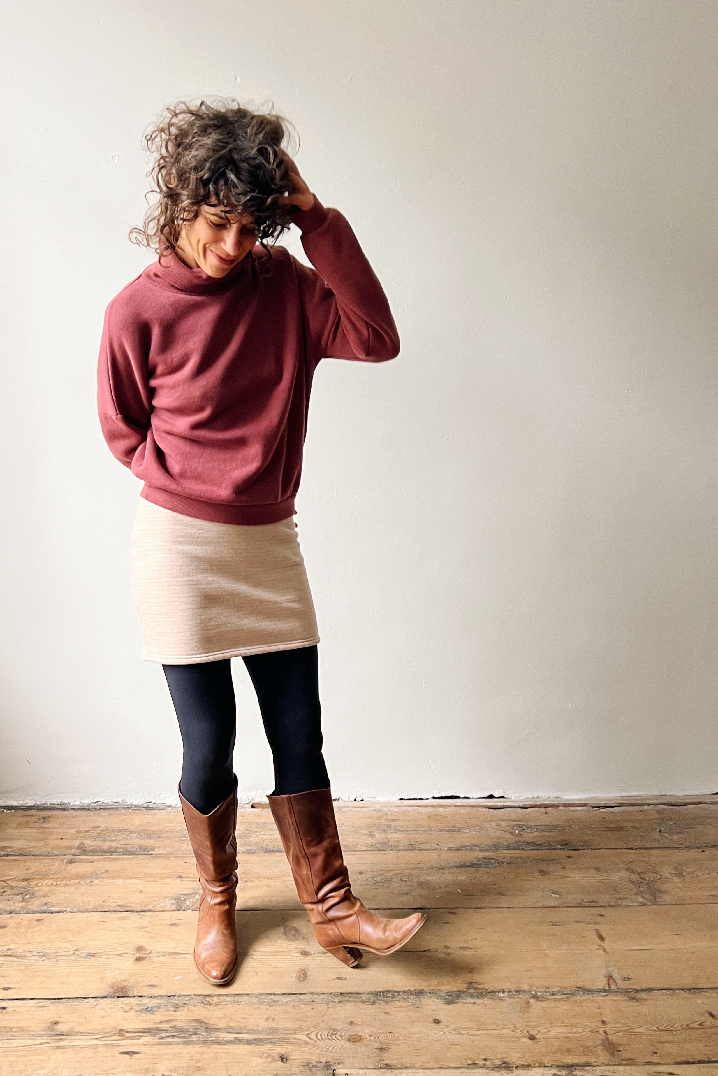 Woman wearing a figue sweater, beige skirt, and brown knee-high boots against a plain wall.