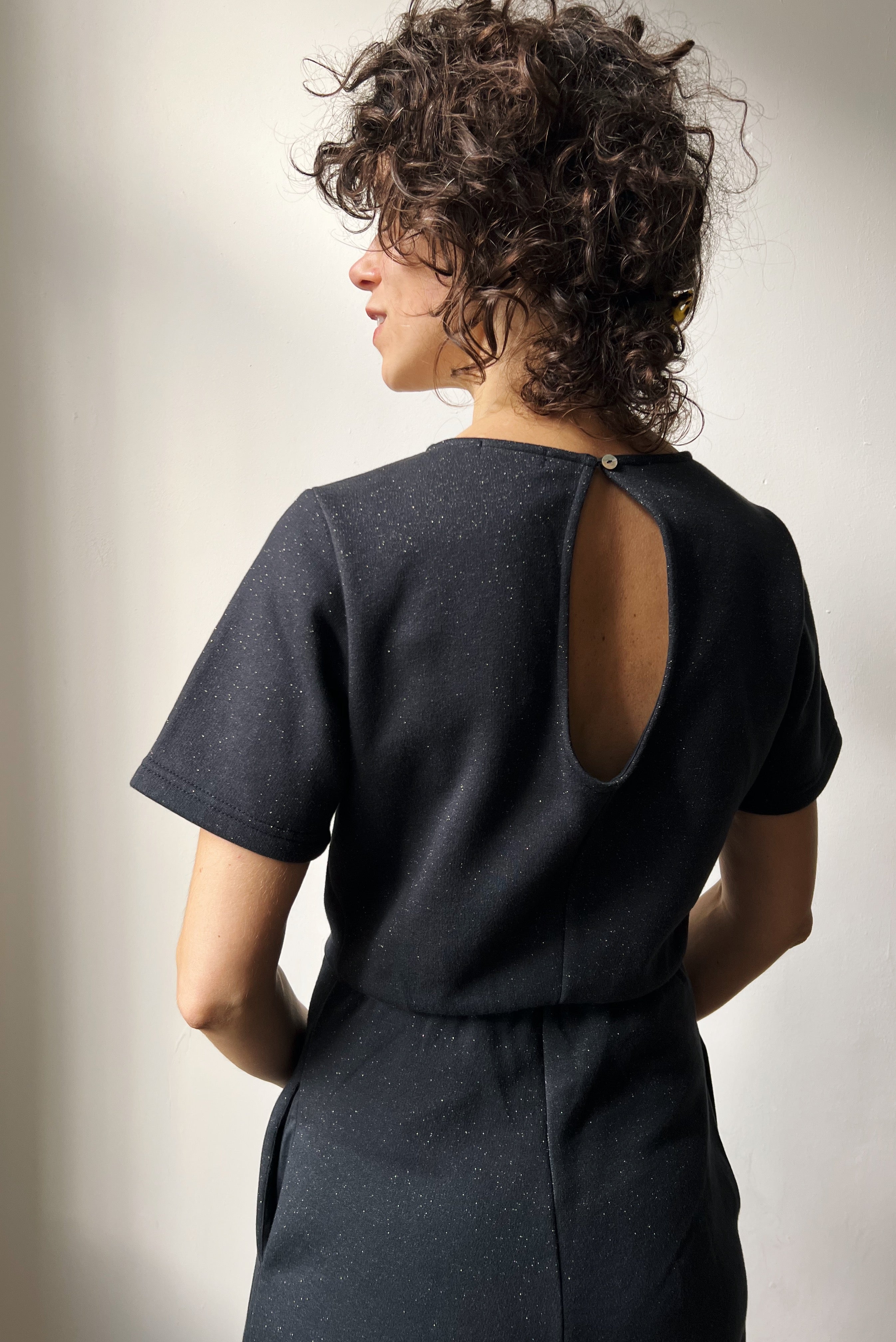 Woman wearing a dark gray sparkly jumpsuit with cutout on the back, standing against a plain white wall.