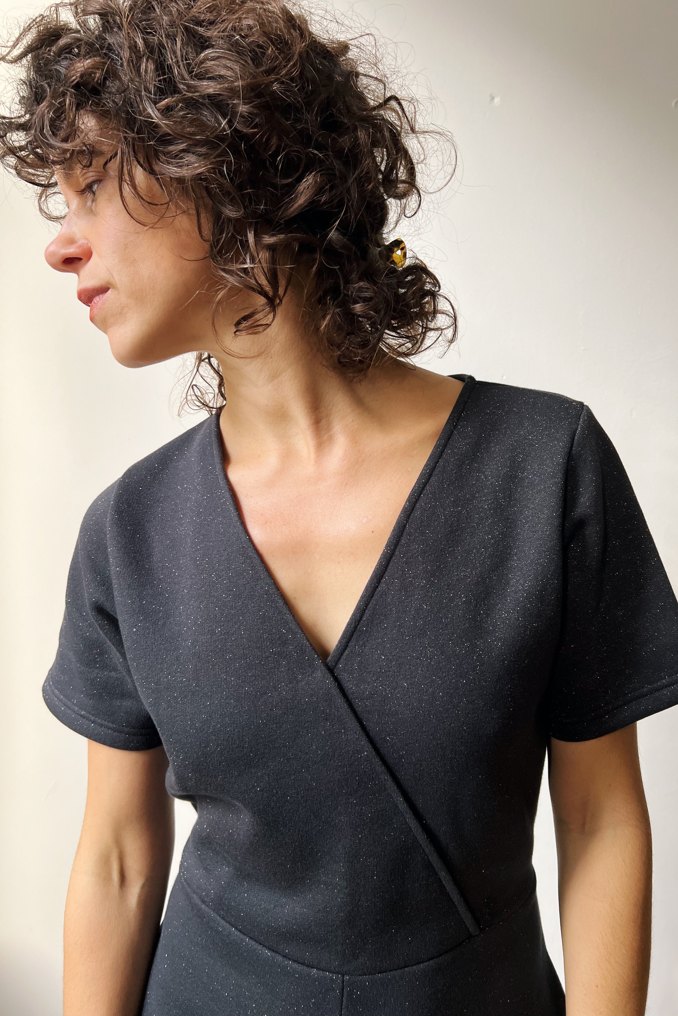 Woman wearing a dark gray sparkly jumpsuit standing against a plain white wall.