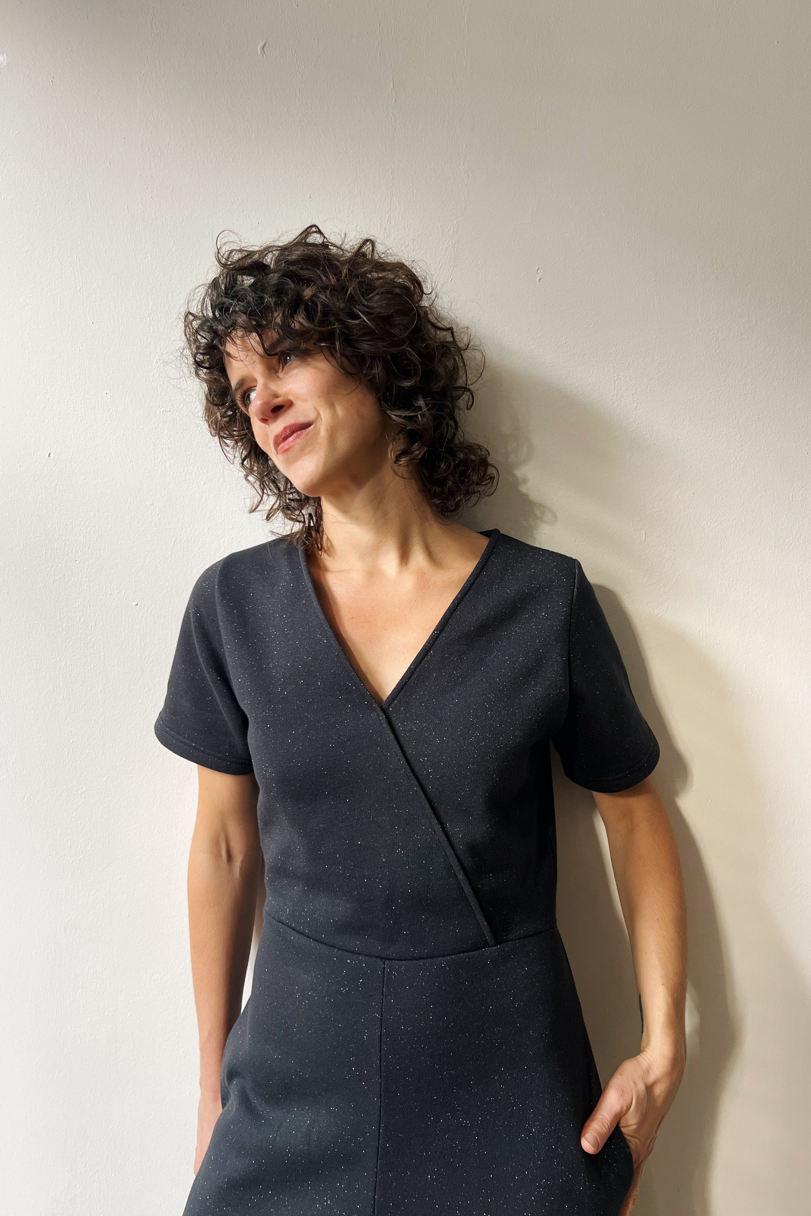 Woman wearing a dark gray sparkly jumpsuit standing against a plain white wall.