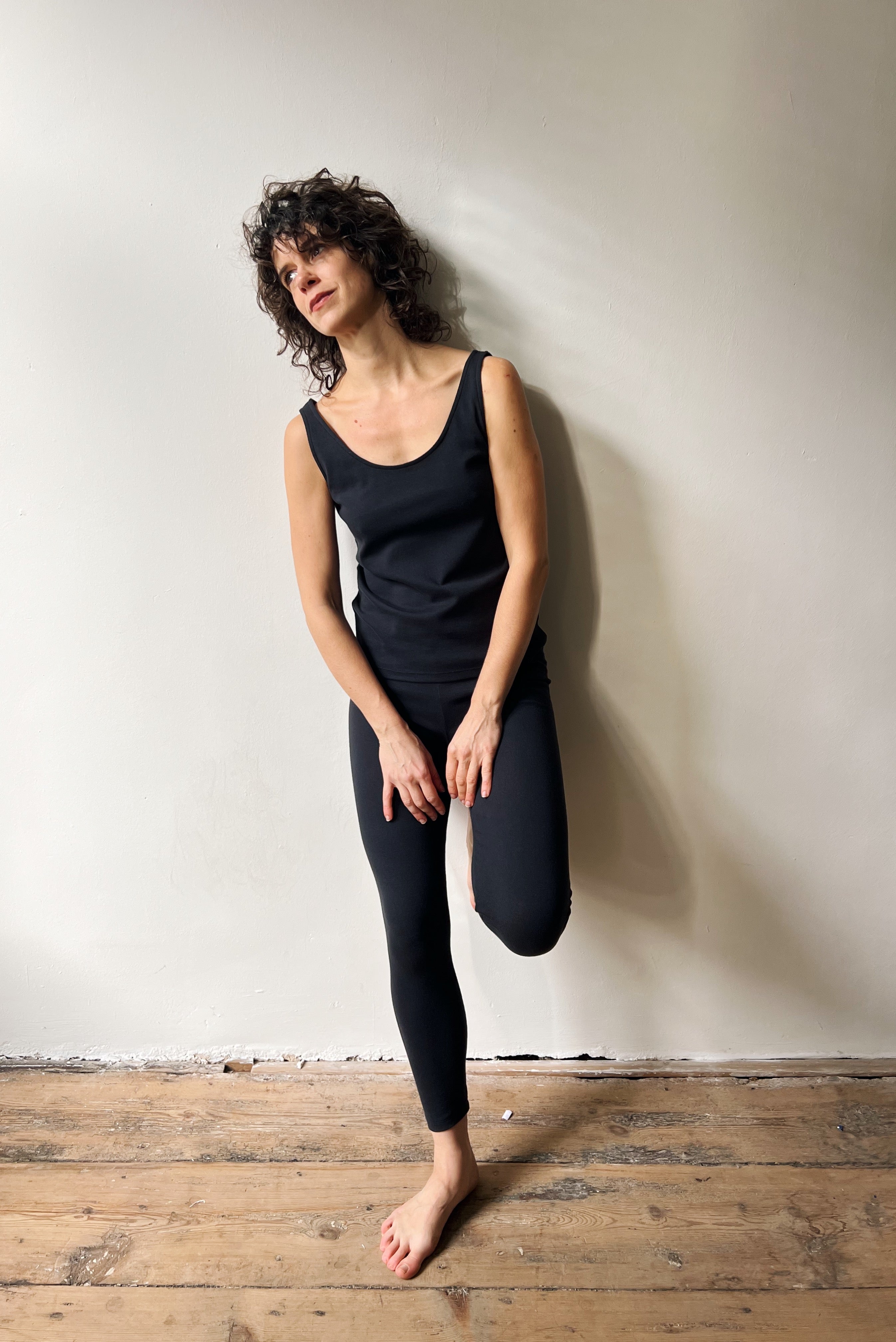 Woman in black tank top and leggings standing against a white wall on a wooden floor.