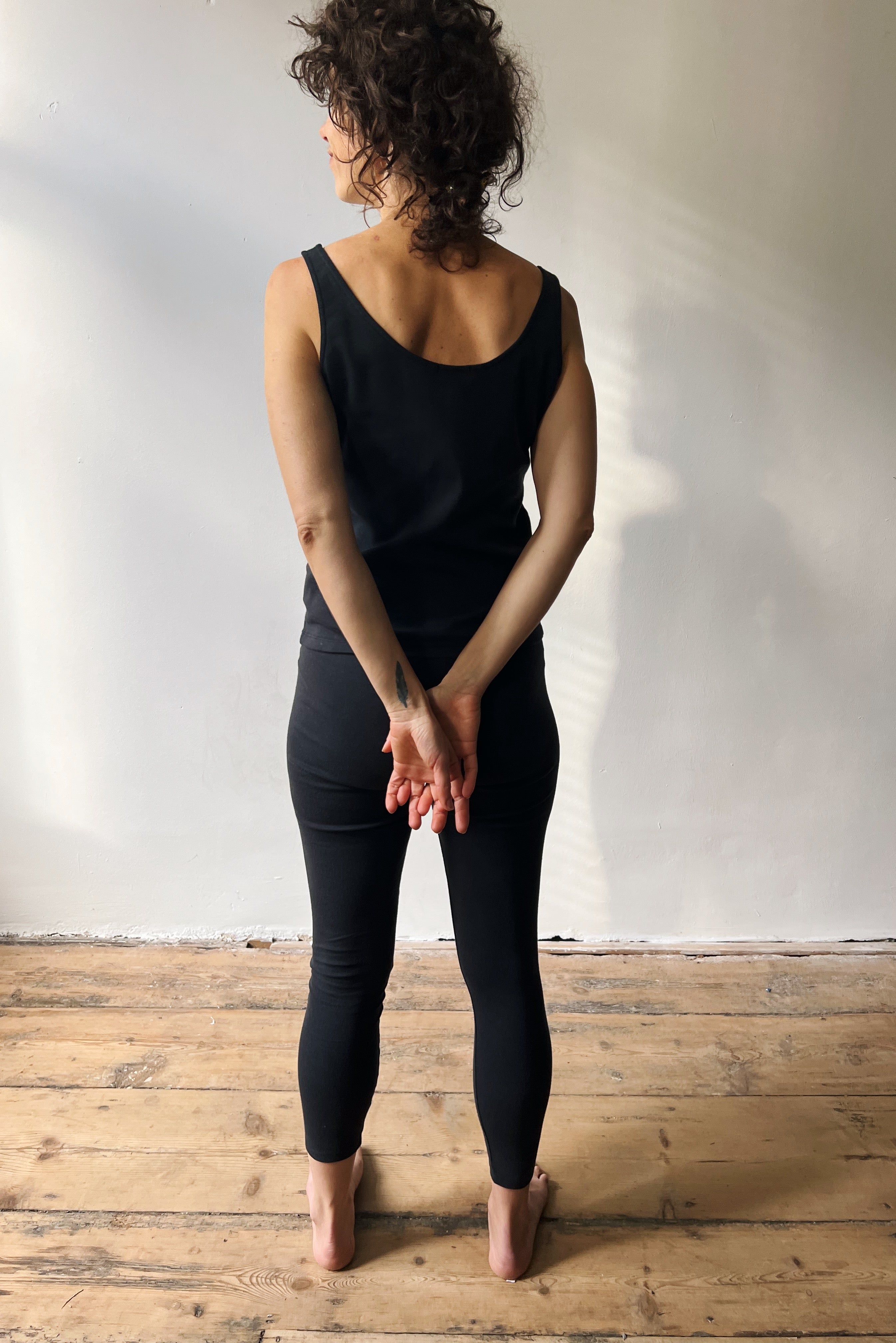 Person wearing a black sleeveless top and leggings standing on a wooden floor with a white wall background.