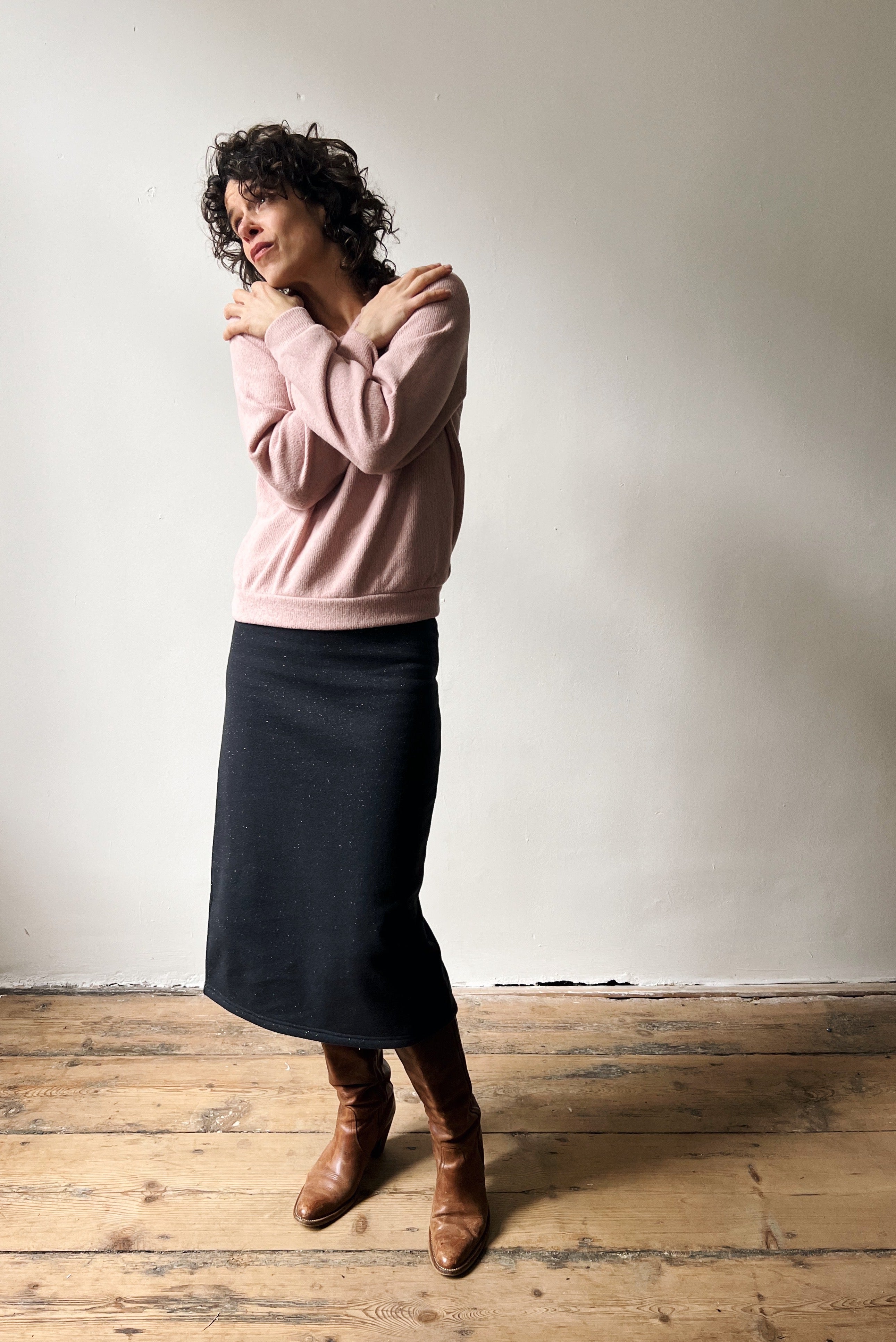 Woman wearing a pink sweater and sparkly dark gray skirt standing against a plain wall.