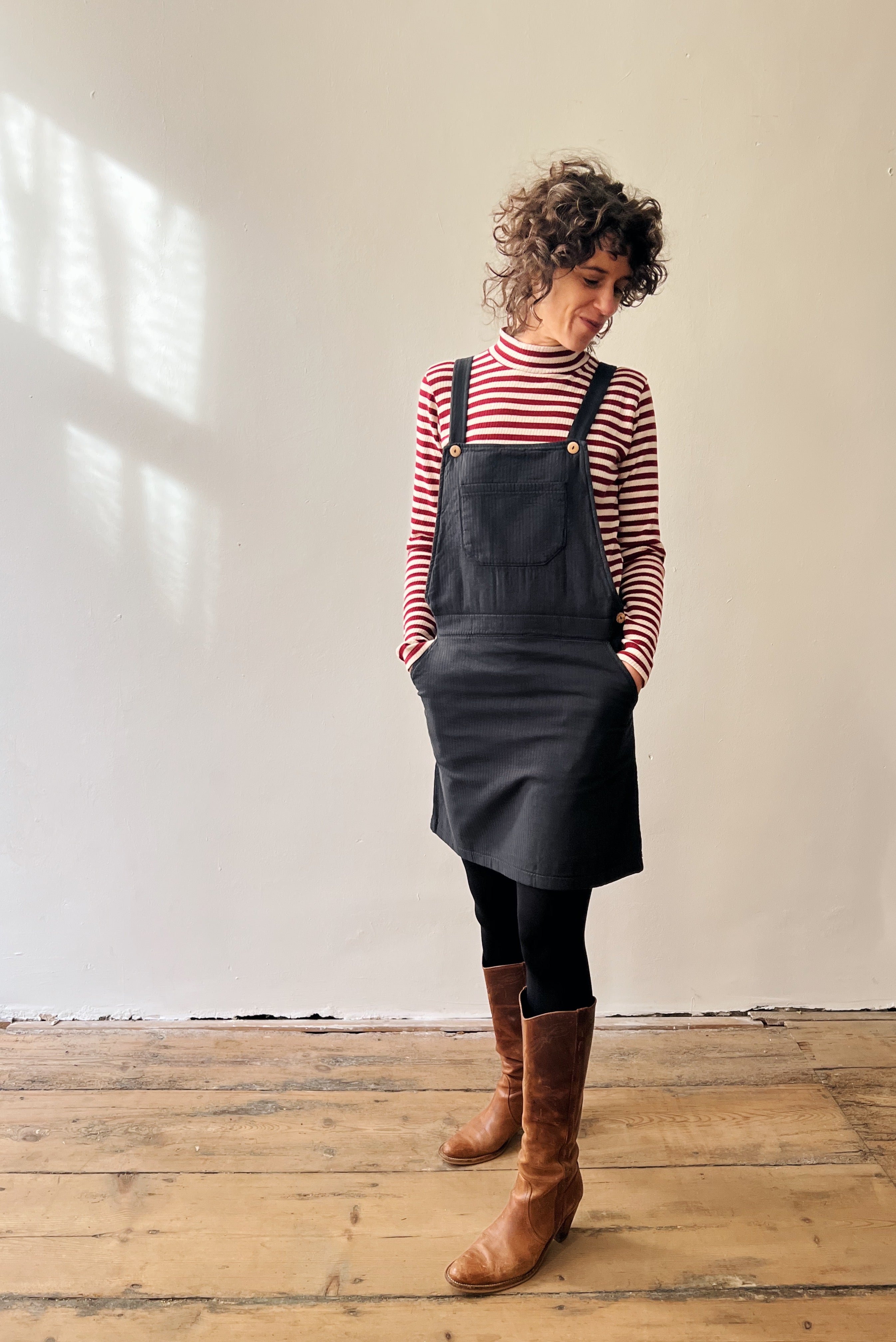 Woman wearing a striped shirt, antra salopettedress, and brown boots in a room with wooden flooring and white walls.