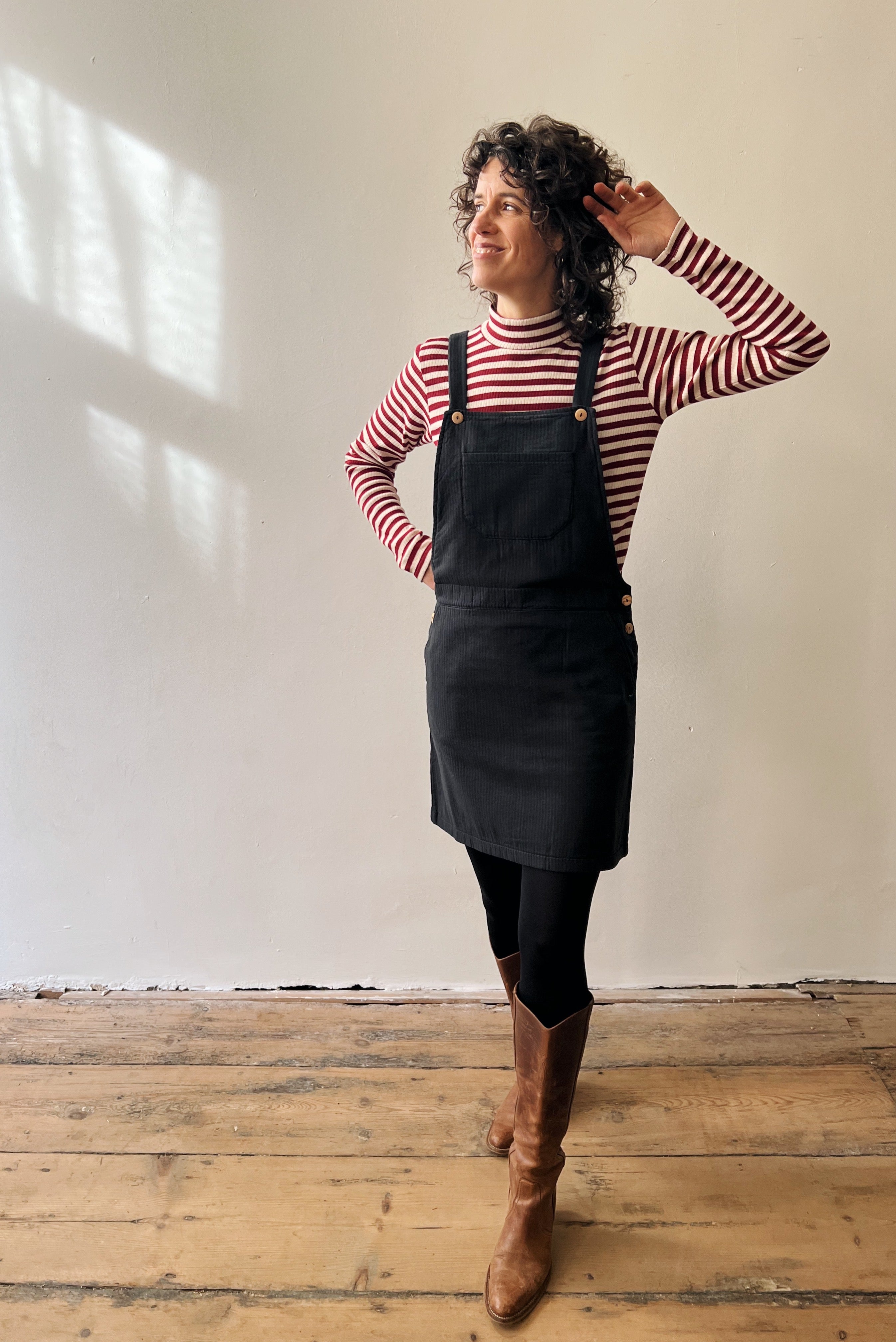 Woman wearing a striped shirt, antra salopettedress, and brown boots in a room with wooden flooring and white walls.