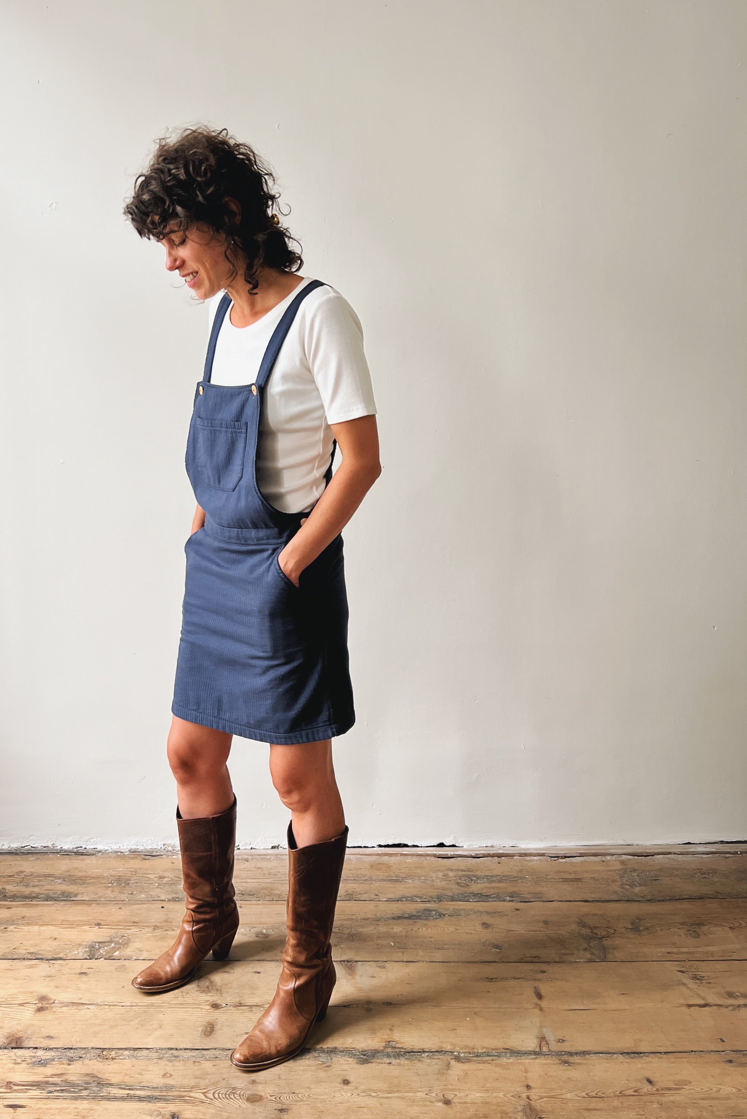 Person wearing a white shirt and blue corduroy salopettedress with kneeboots against a plain background