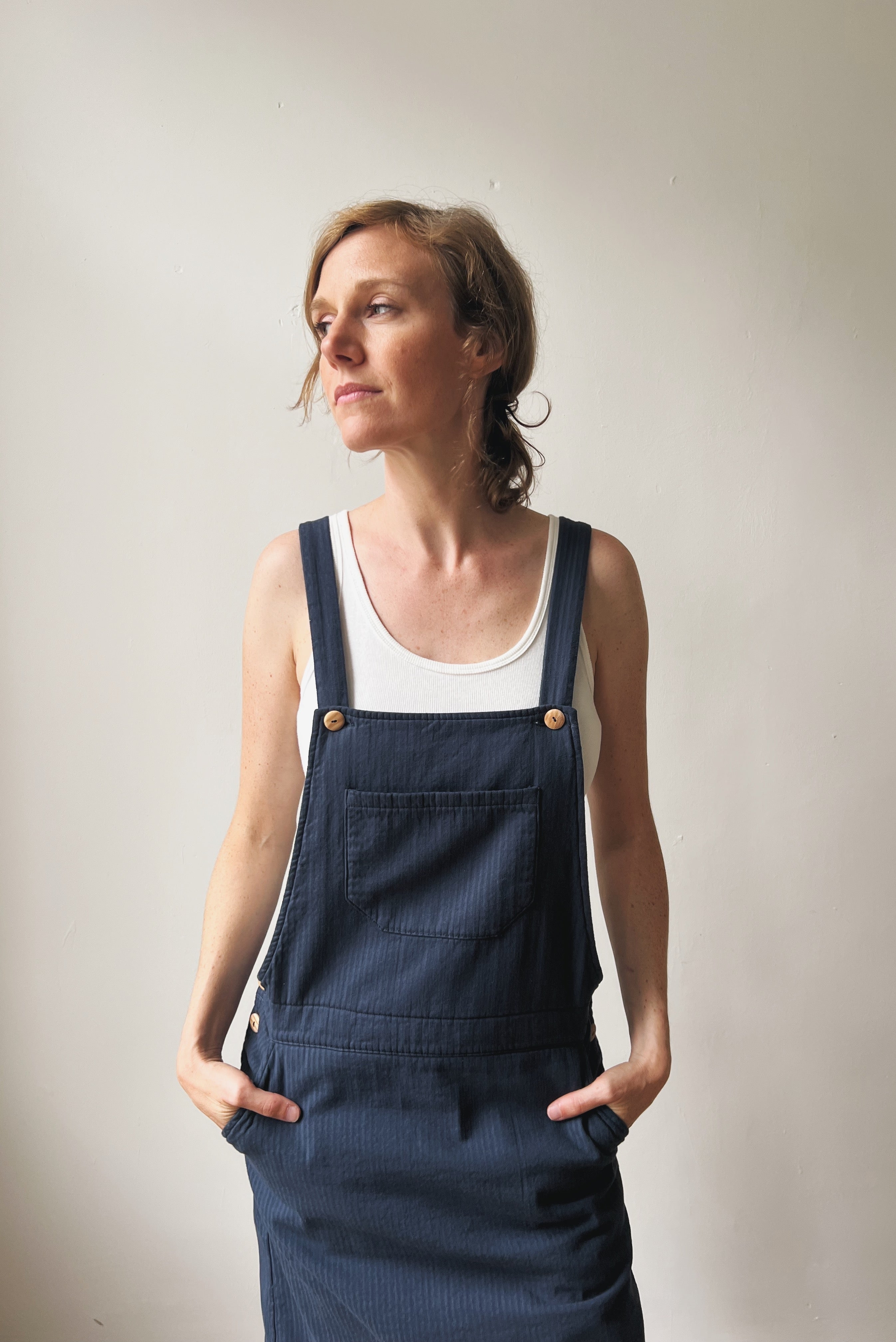 Woman wearing a navy blue corduroy salopette dress over a white tank top against a plain background