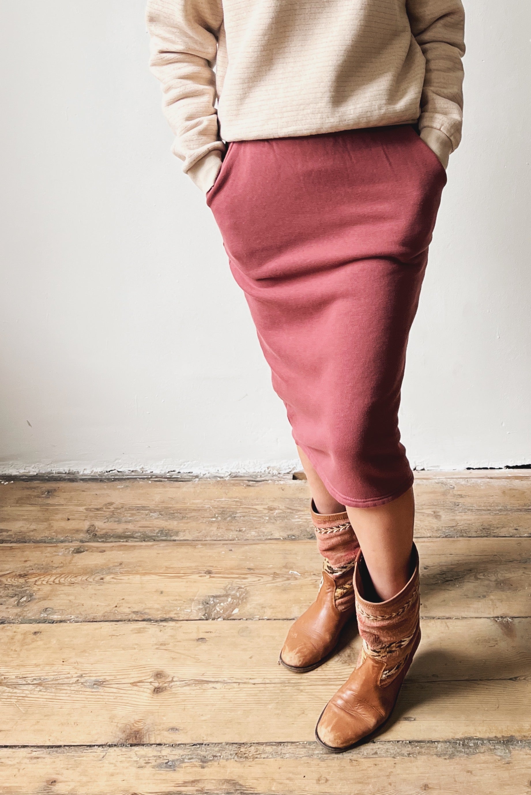 Person wearing a beige sweater, figue skirt, and brown boots on a wooden floor.