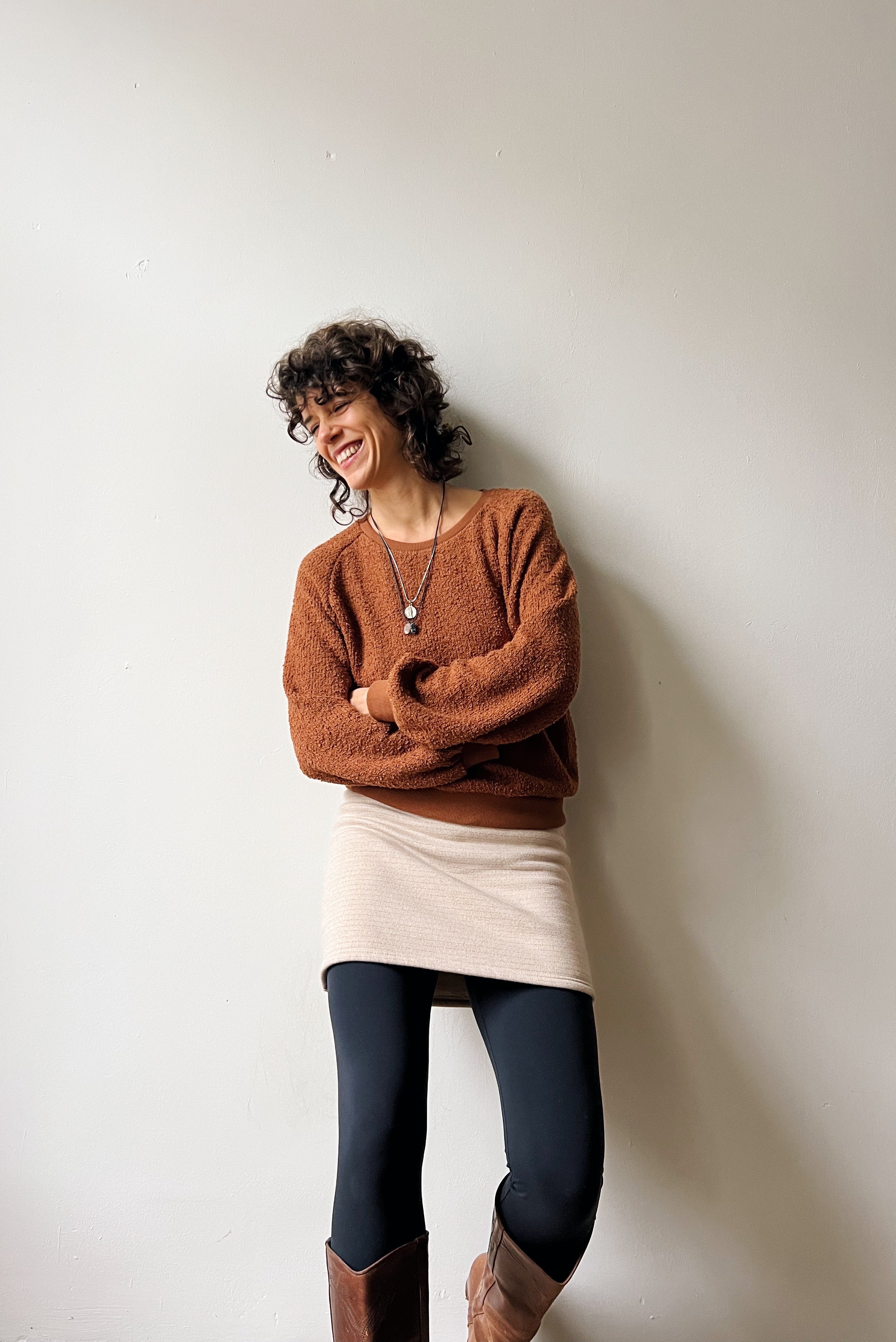 Woman wearing a brown teddy sweater and beige skirt with subtle bronze stripes against a white wall
