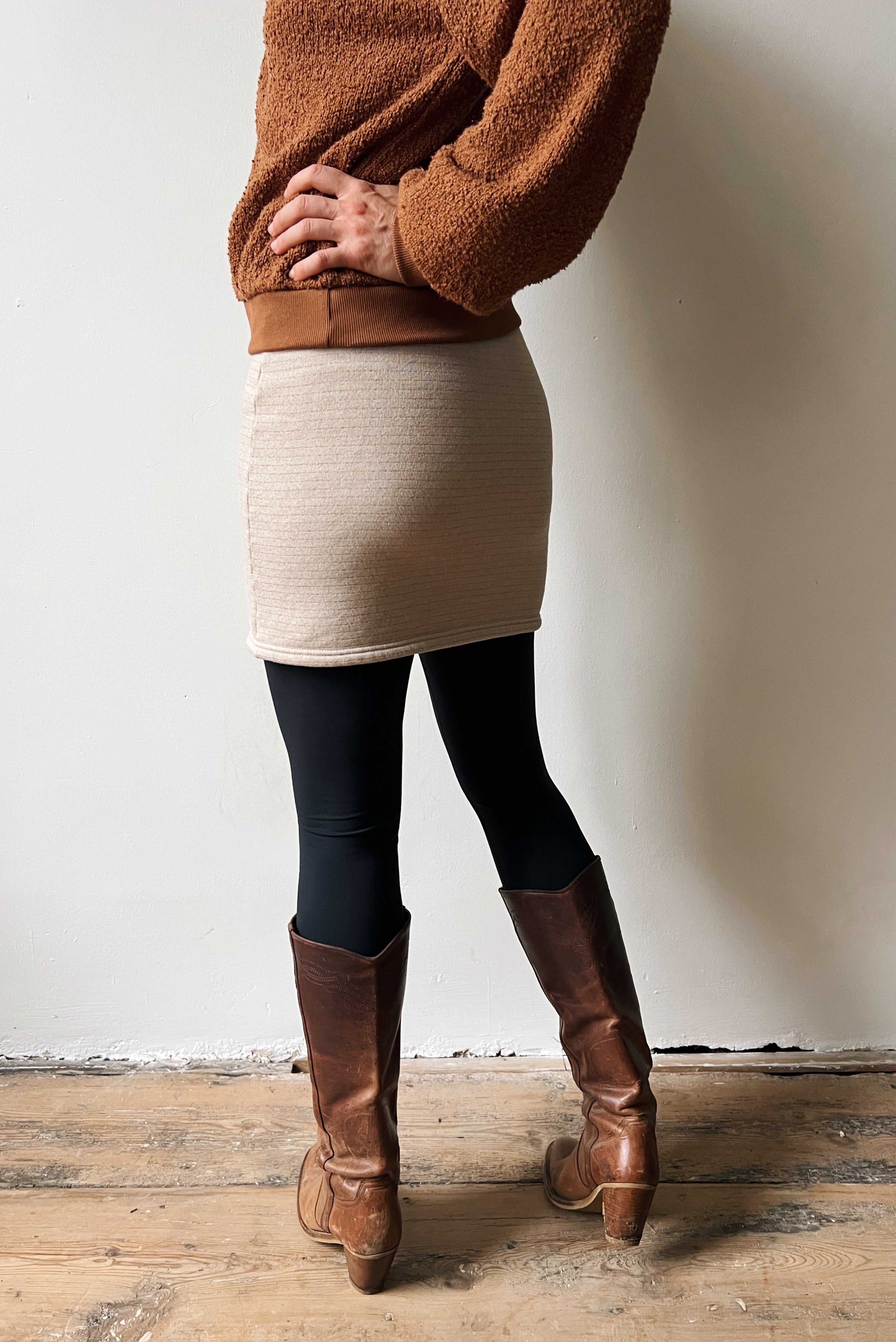 Person wearing a brown sweater, beige skirt with subtle bronze stripes and brown boots against a white wall.