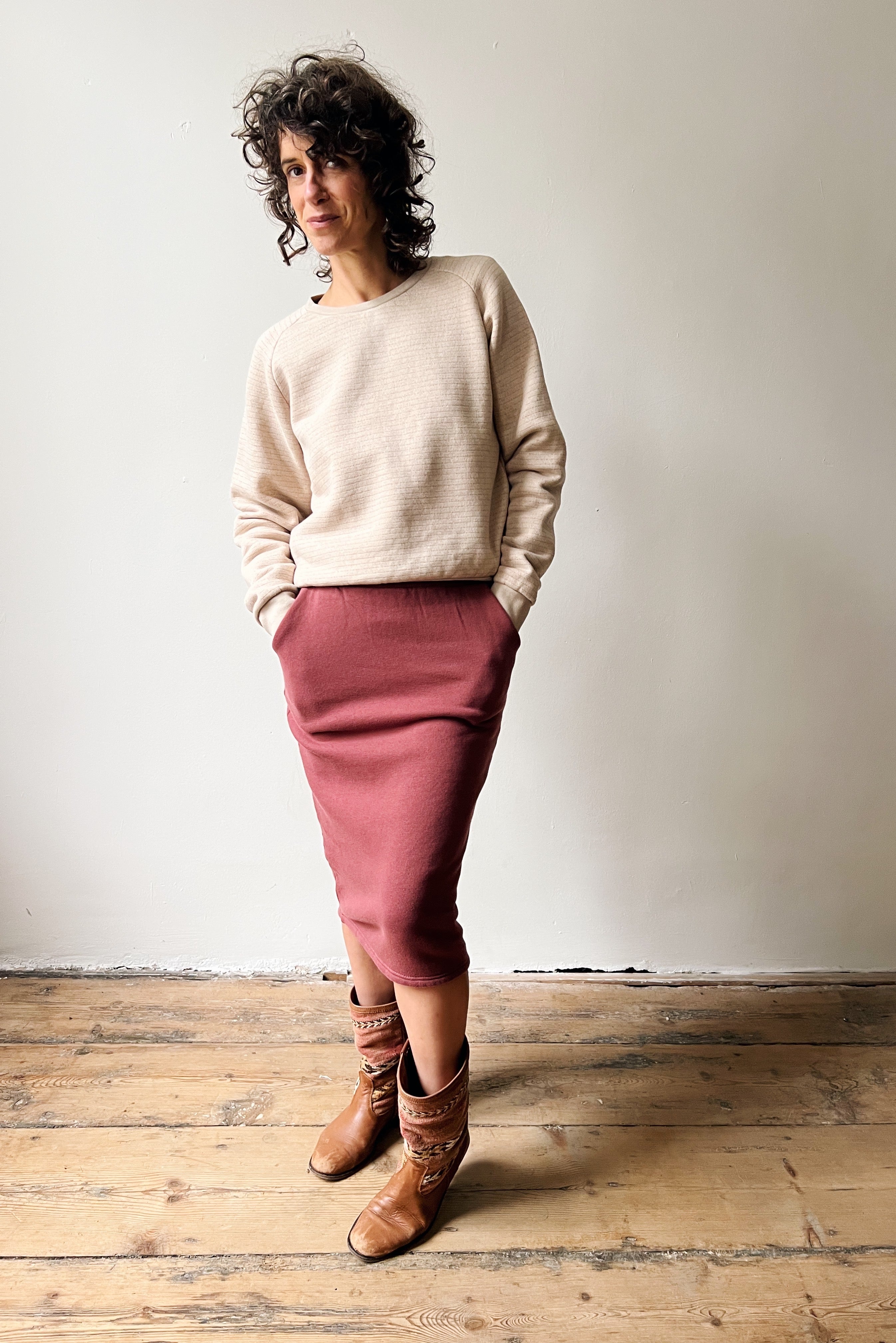 Woman wearing a beige sweater and figue skirt standing on a wooden floor with a white wall background