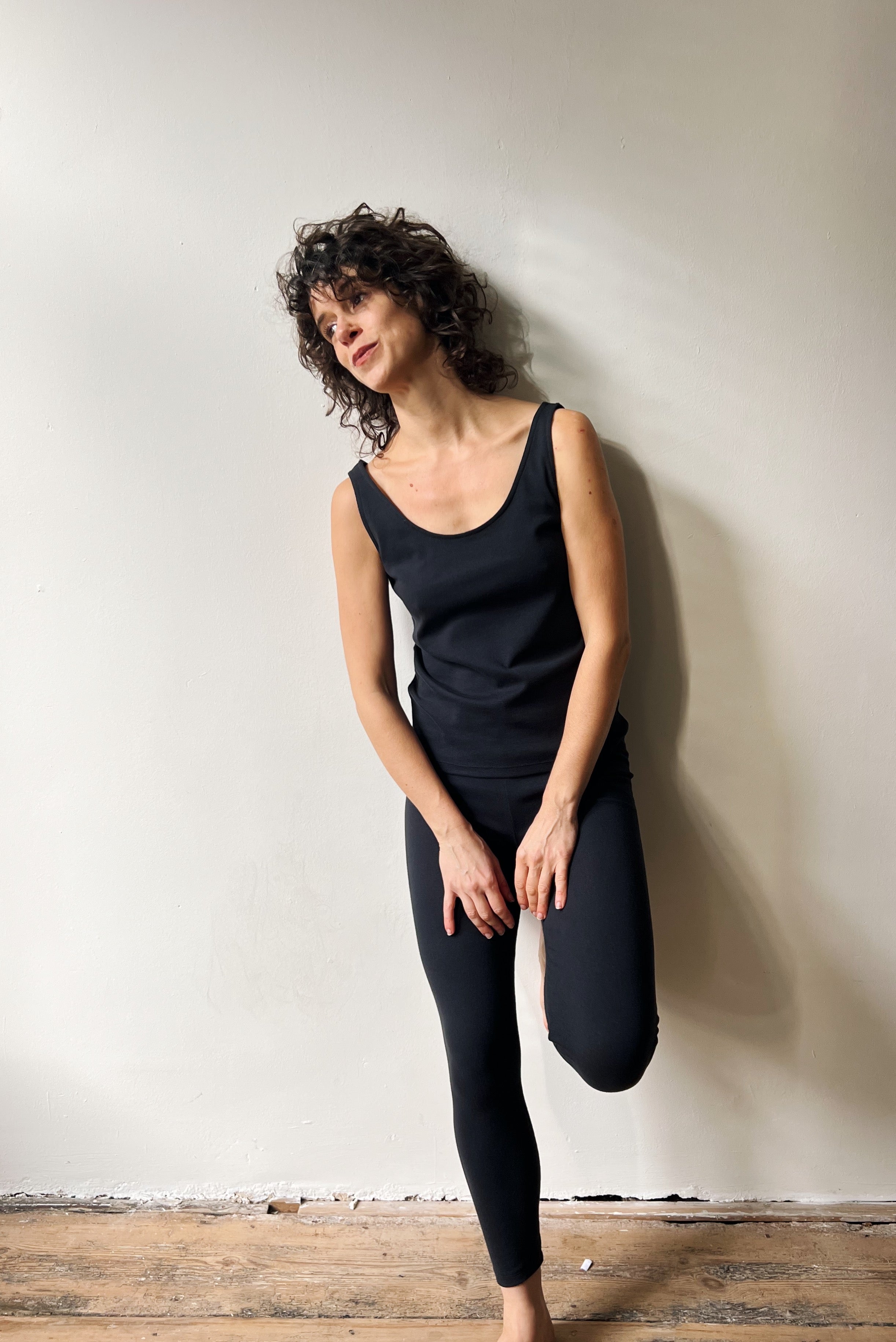 Woman wearing a black sleeveless top and leggings against a plain wall.