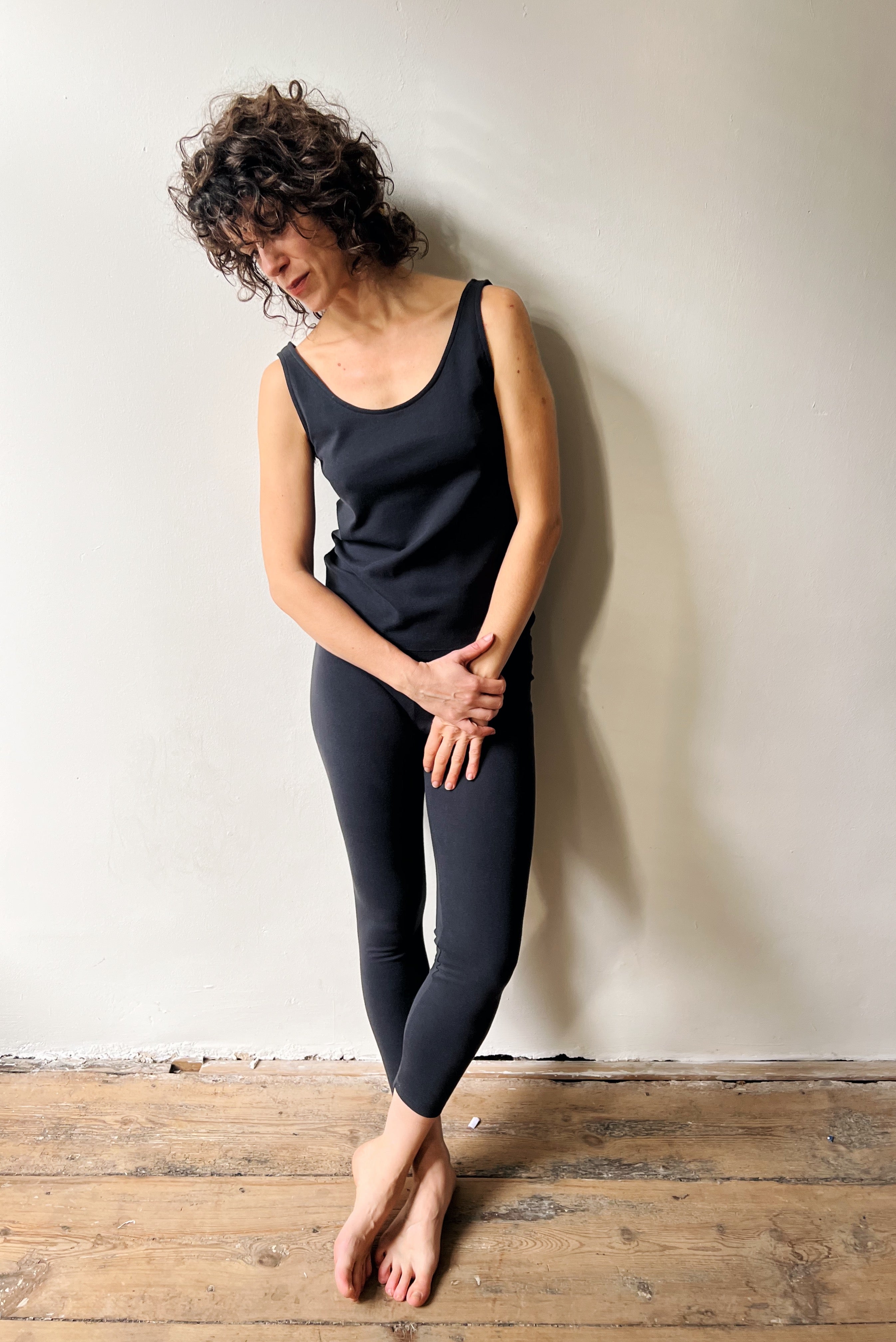 Woman wearing a black tank top and leggings against a white wall.