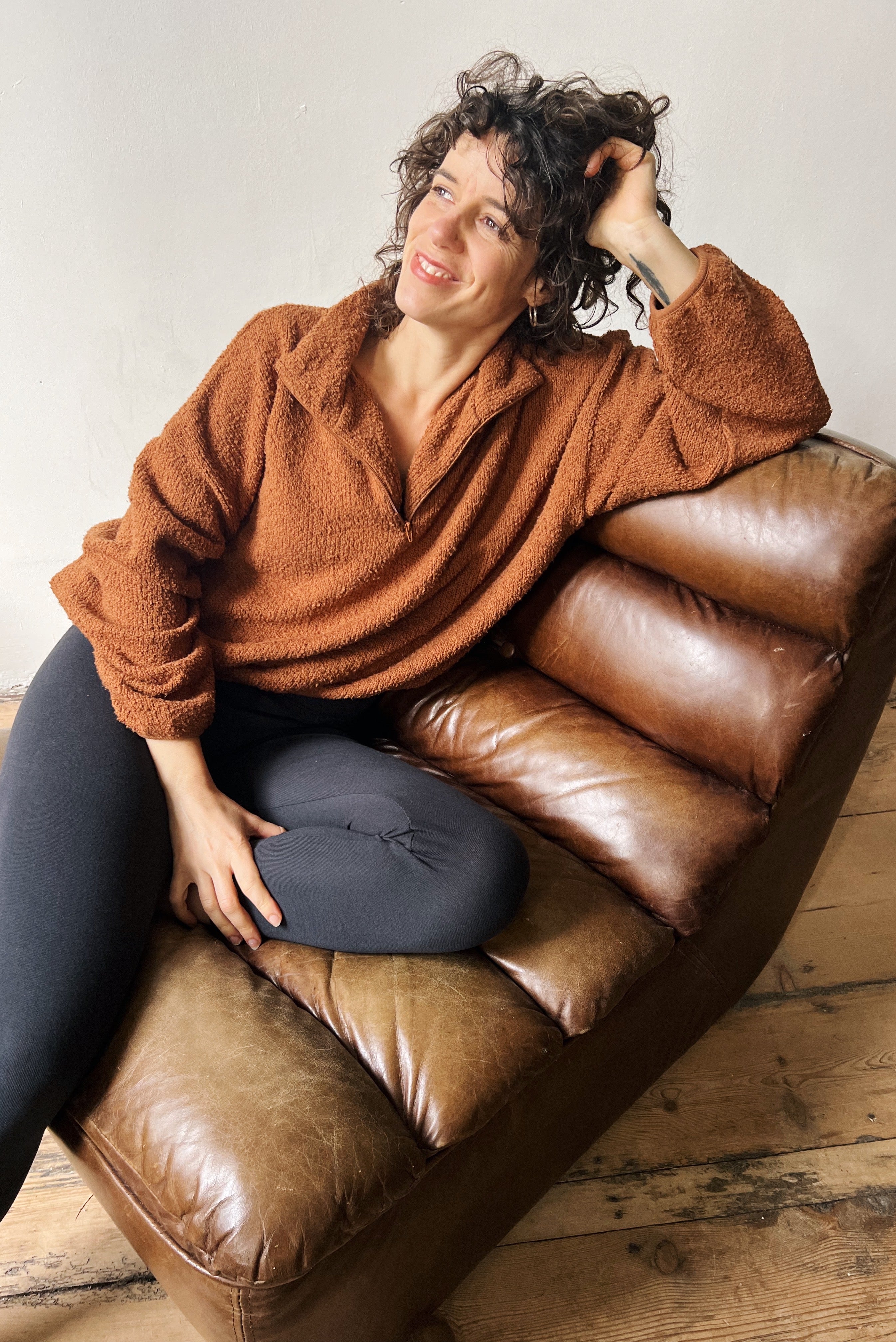 Woman sitting on a brown leather couch with a brown teddy sweater and black leggings on.