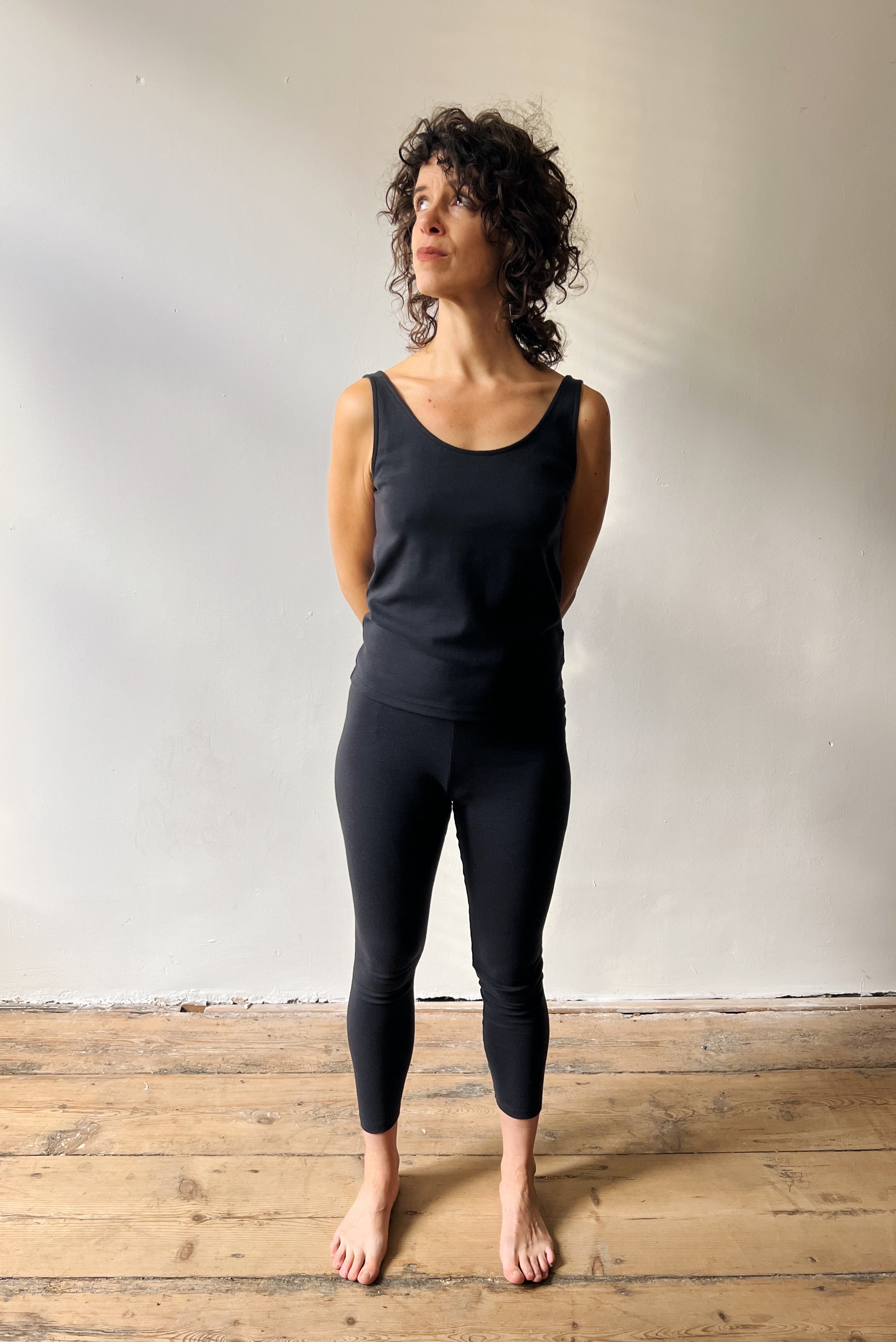 Woman wearing a black tank top and leggings standing on a wooden floor against a white wall.