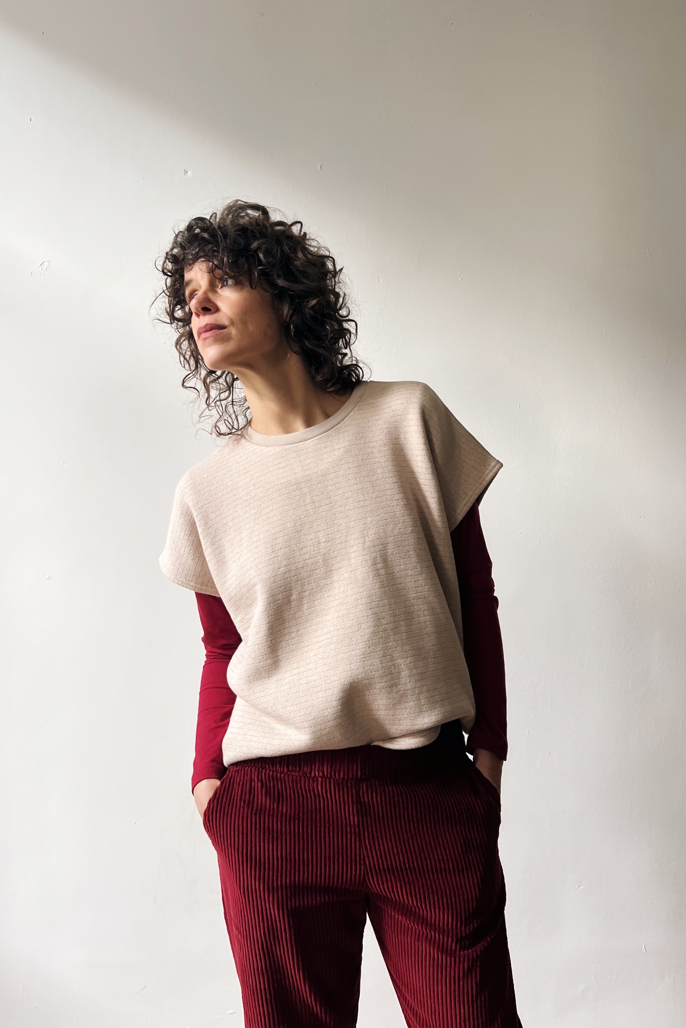 Woman wearing a beige shortsleeve sweater, red longsleeve and red pants standing against a white wall.