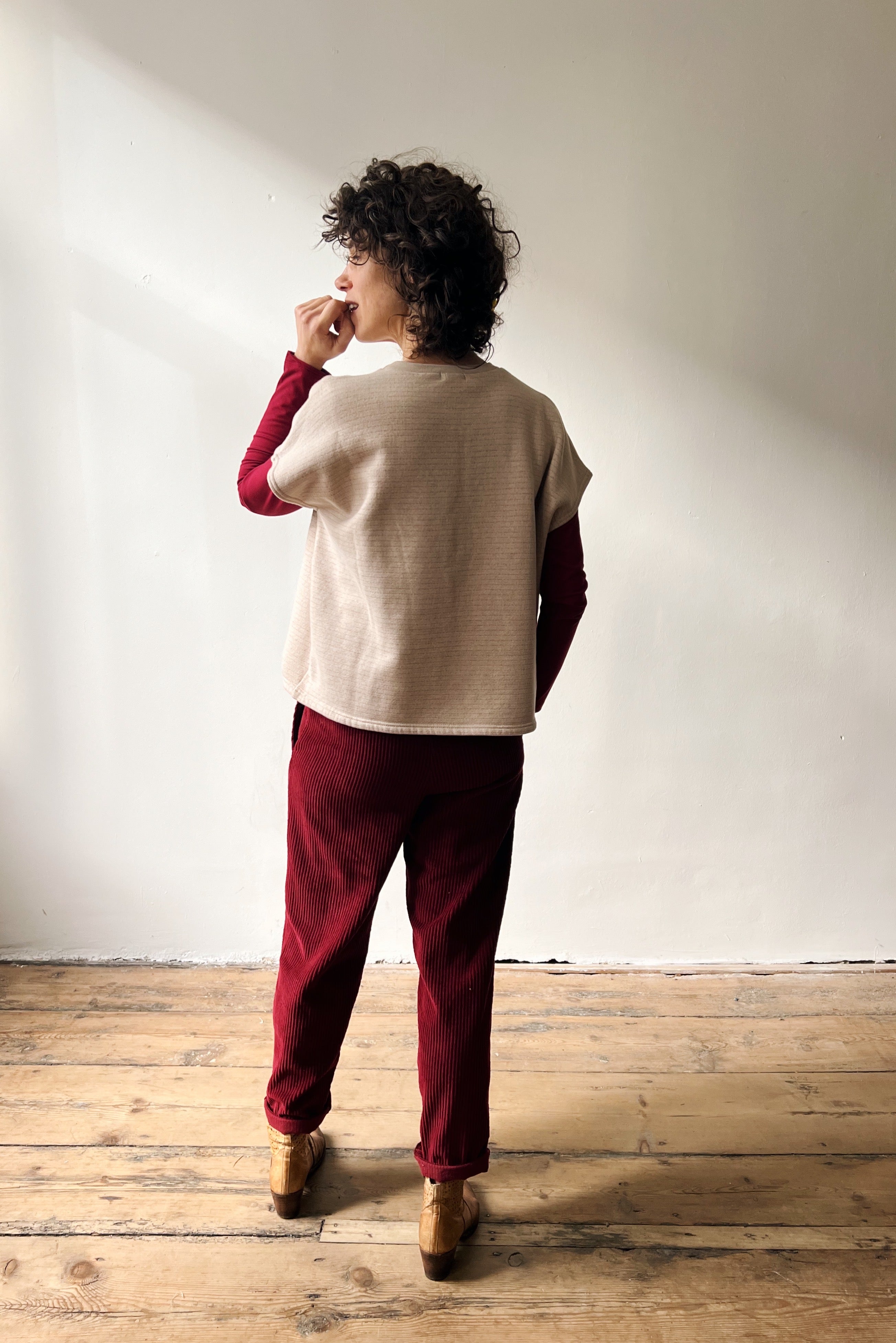 Woman wearing a beige shortsleeve sweater, red longsleeve and red pants standing against a white wall.