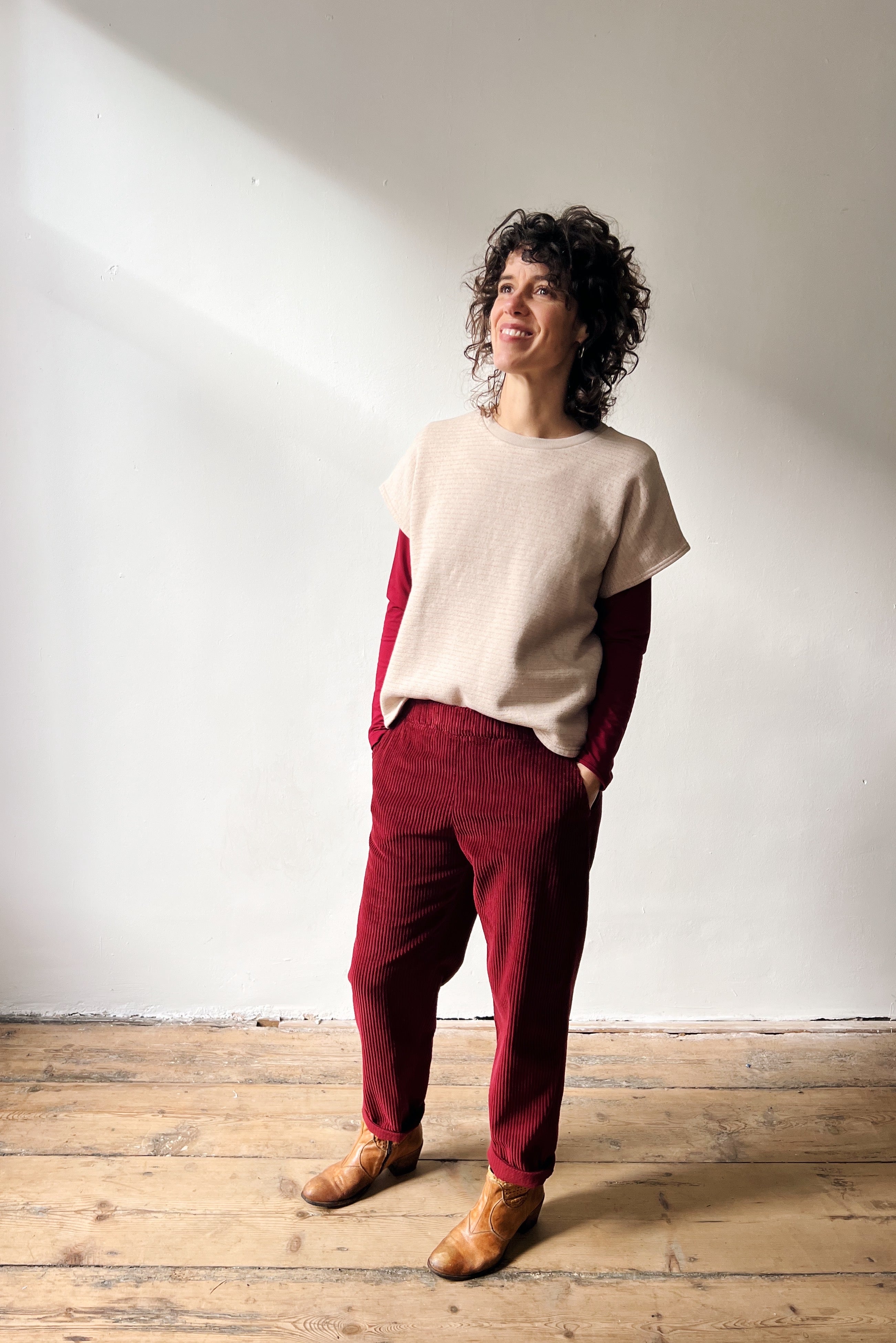 Woman wearing a beige shortsleeve sweater, red longsleeve and red pants standing against a white wall.